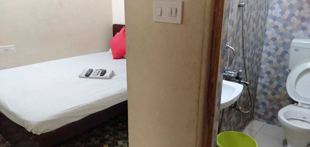 A small room with a bed, a bedside table with a remote control, a pink pillow, a bathroom with a toilet, a sink, and a shower.