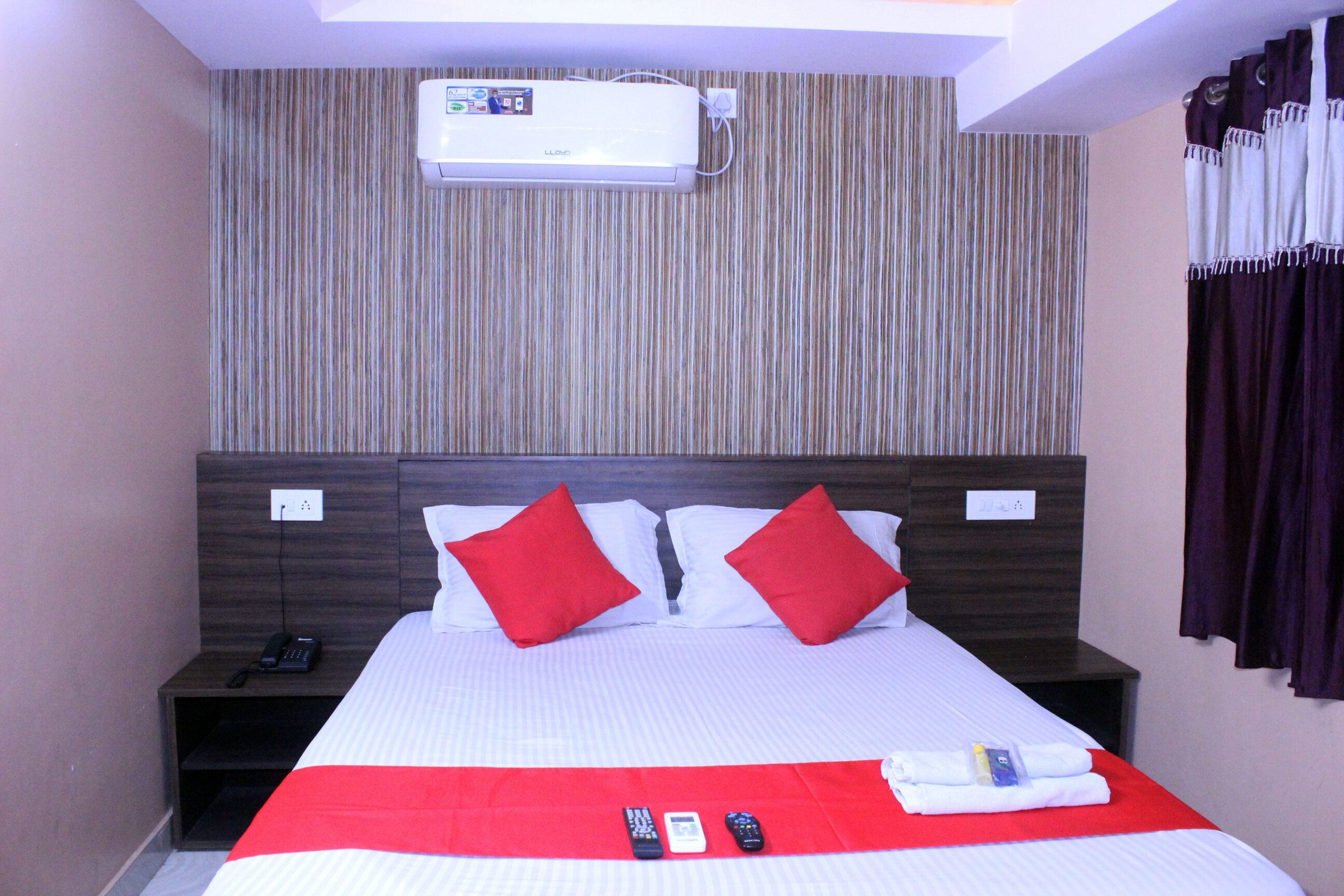 Habitación de hotel con cama, almohadas rojas, colcha roja, aire acondicionado blanco y cabecero de madera oscura.