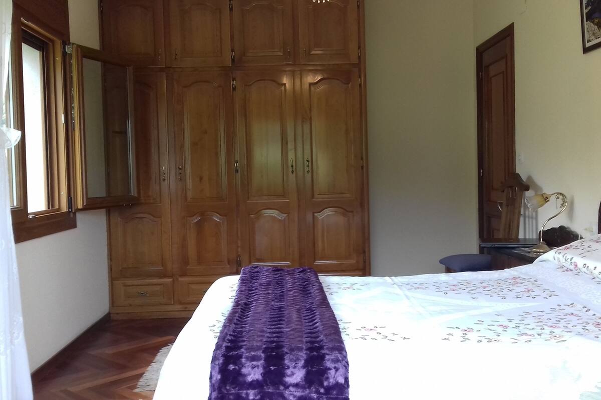 Quarto com guarda-roupa espaçoso, cama com cobertor roxo e mesa de cabeceira com abajur.