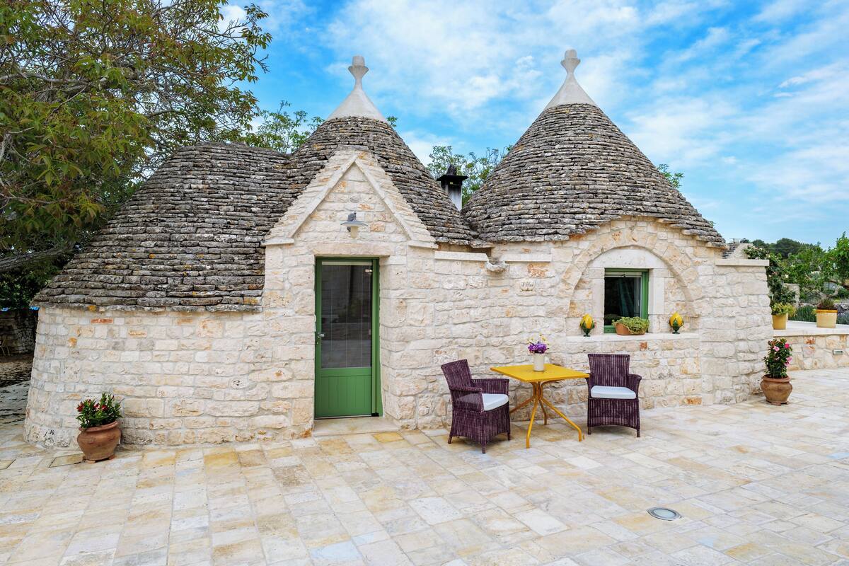 Un edificio de piedra con techos cónicos, una puerta verde y un pequeño patio con dos sillas de mimbre y una mesa.