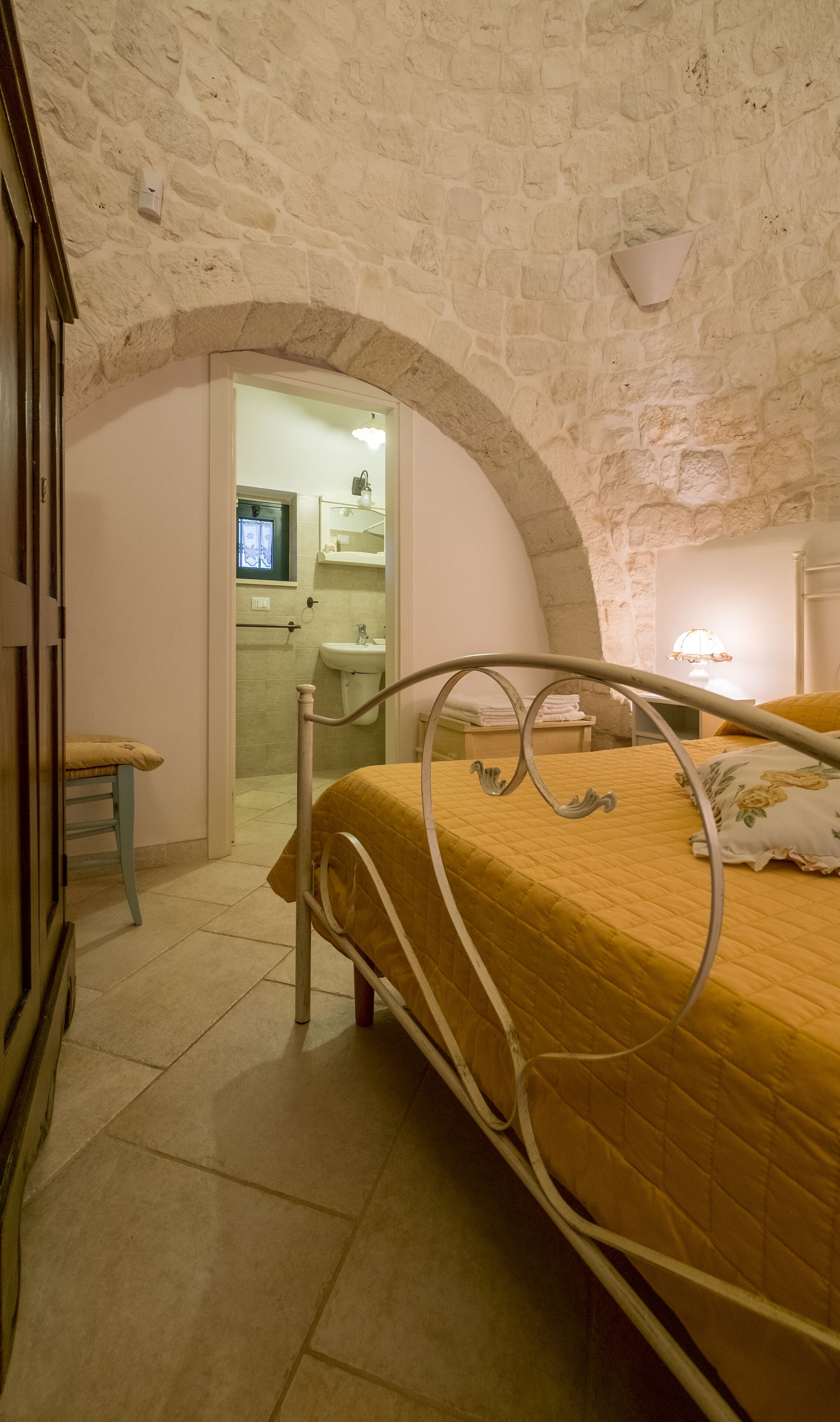 Een slaapkamer met een stenen muur, een bed met metalen frame, een houten stoel en een badkamer zichtbaar door een boog.