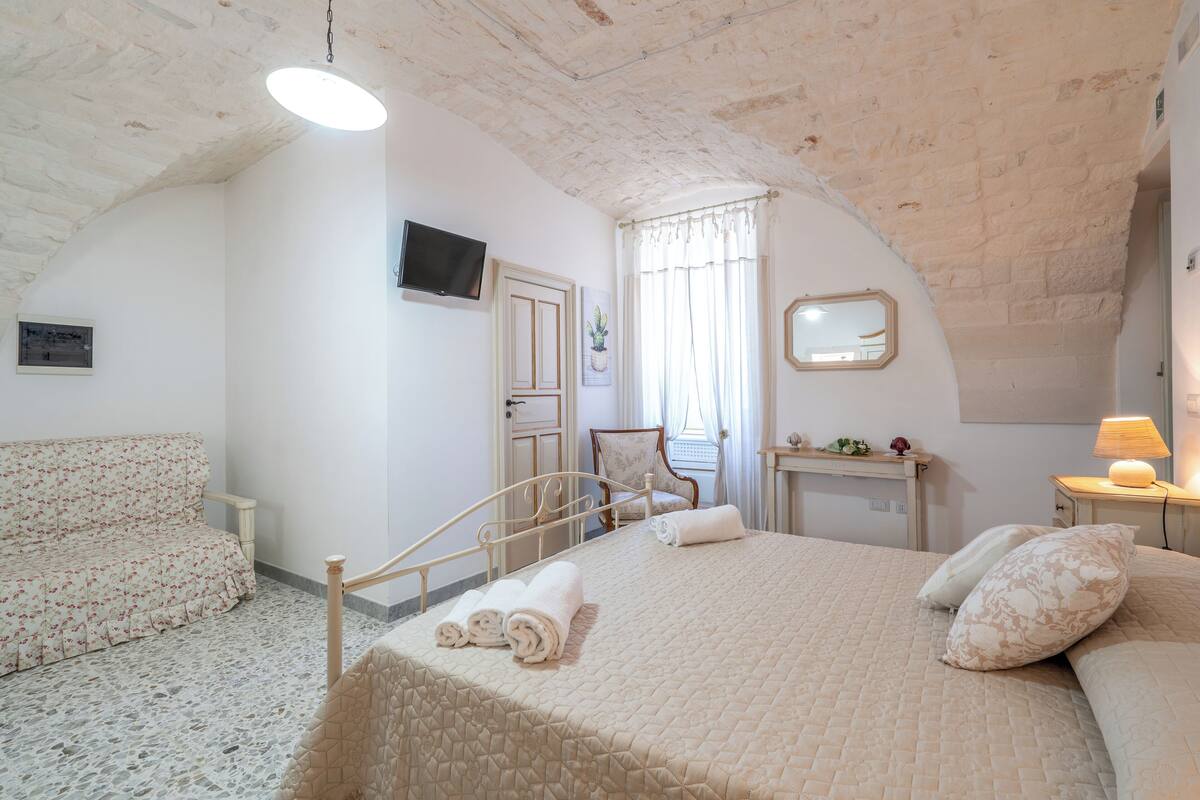 Un dormitorio con cama, una silla, un televisor y una mesita de noche con lámpara.