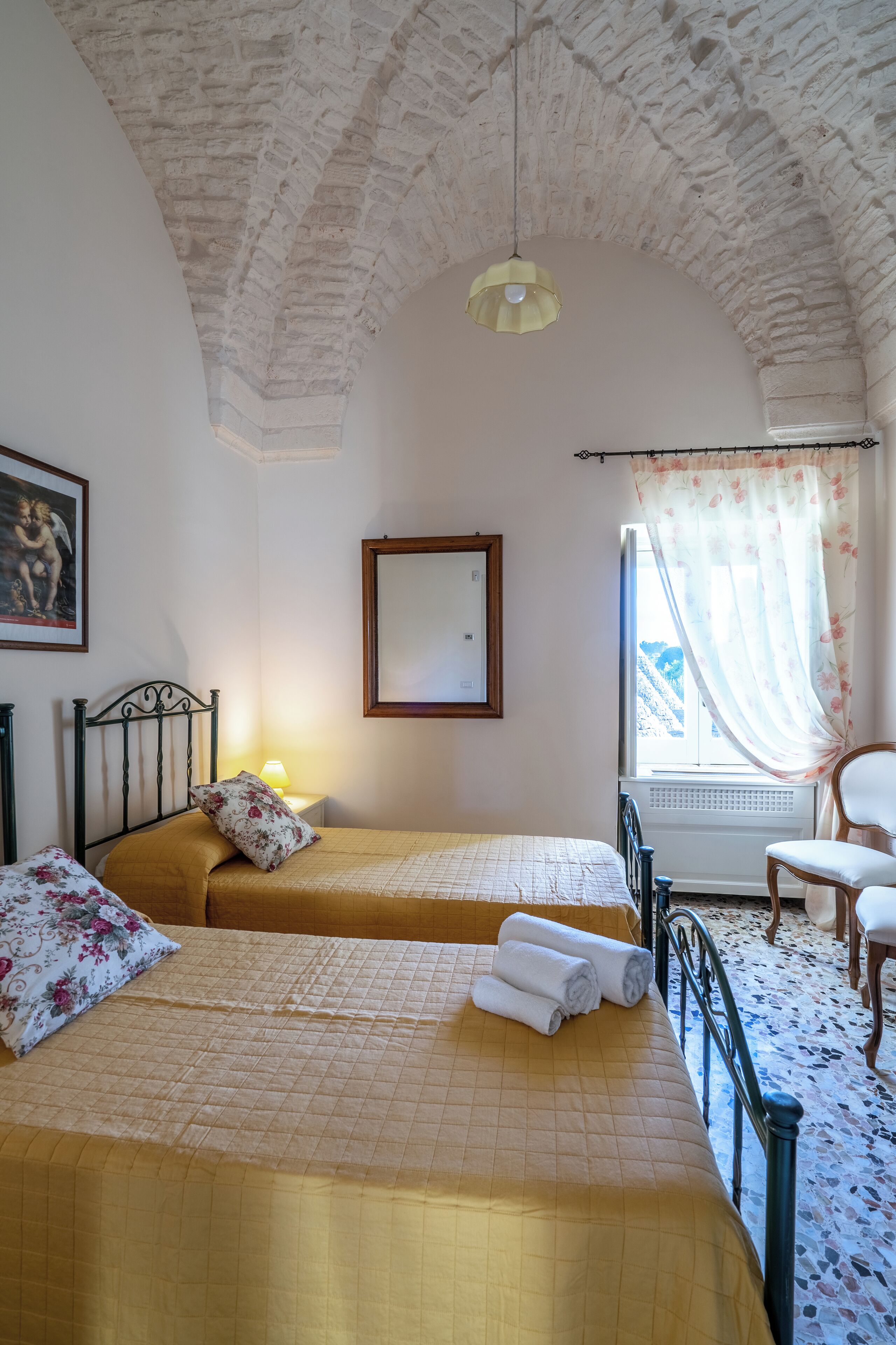 Un dormitorio con dos camas, una ventana con cortinas florales, una silla y un cuadro en la pared.