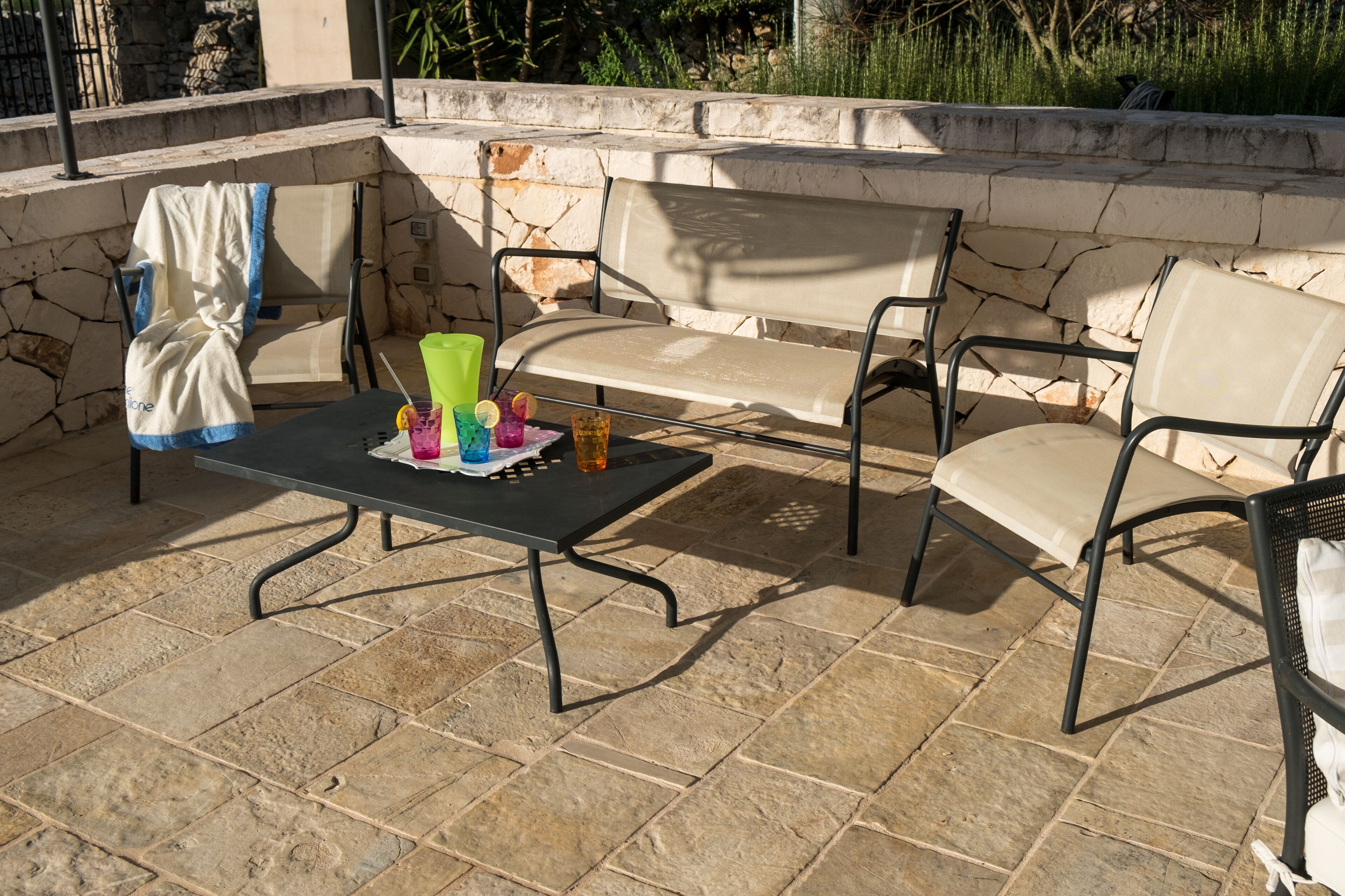 Uma área de pátio externa com piso de pedra, uma mesa com bebidas coloridas e um sofá com uma toalha sobre o apoio de braço.