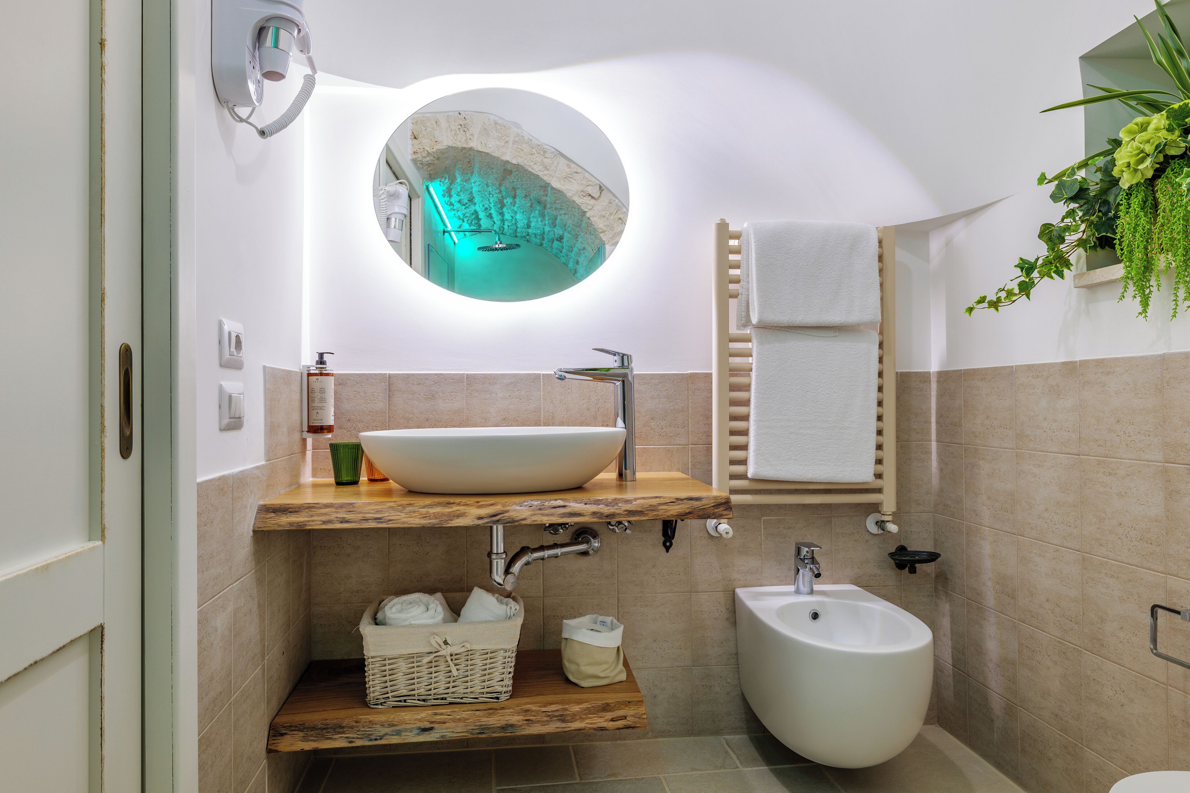 Ein Badezimmer mit rundem Spiegel, Waschbecken in Weiß, einem Bidet, einer Holzablage und einem Handtuchhalter.