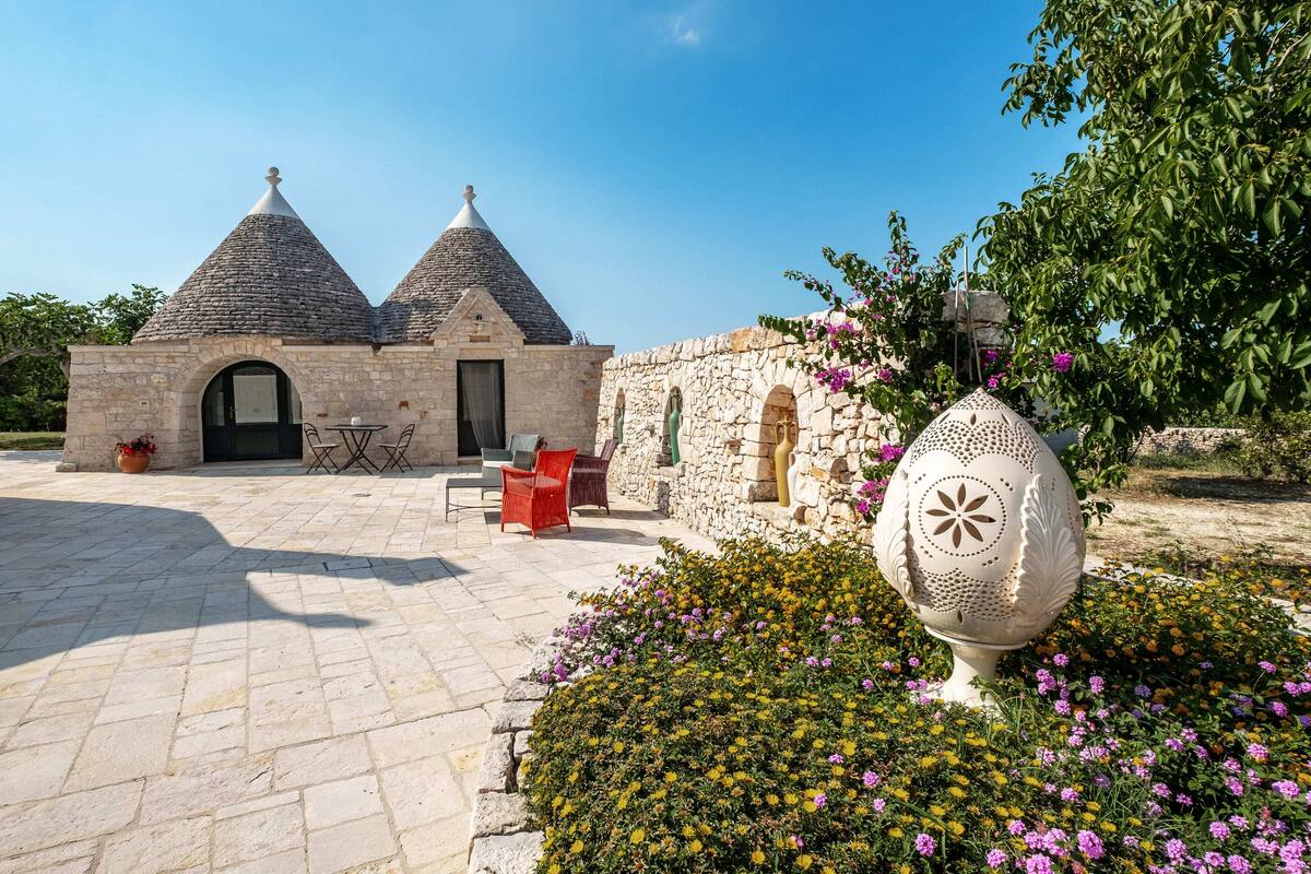 Un edificio de piedra con dos techos cónicos, un pequeño jardín con flores coloridas y un jarro decorativo en primer plano.
