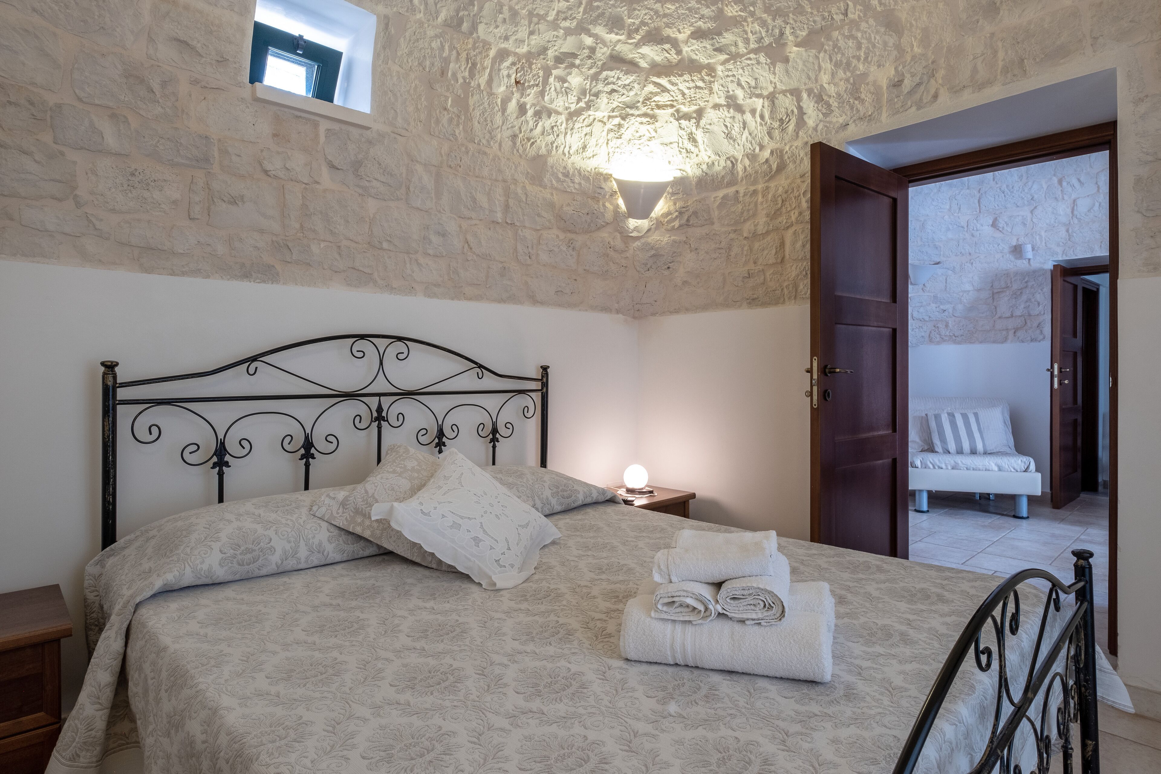 Quarto com parede de pedra, cama com cabeceira metálica, mesa de cabeceira e uma pequena janela.
