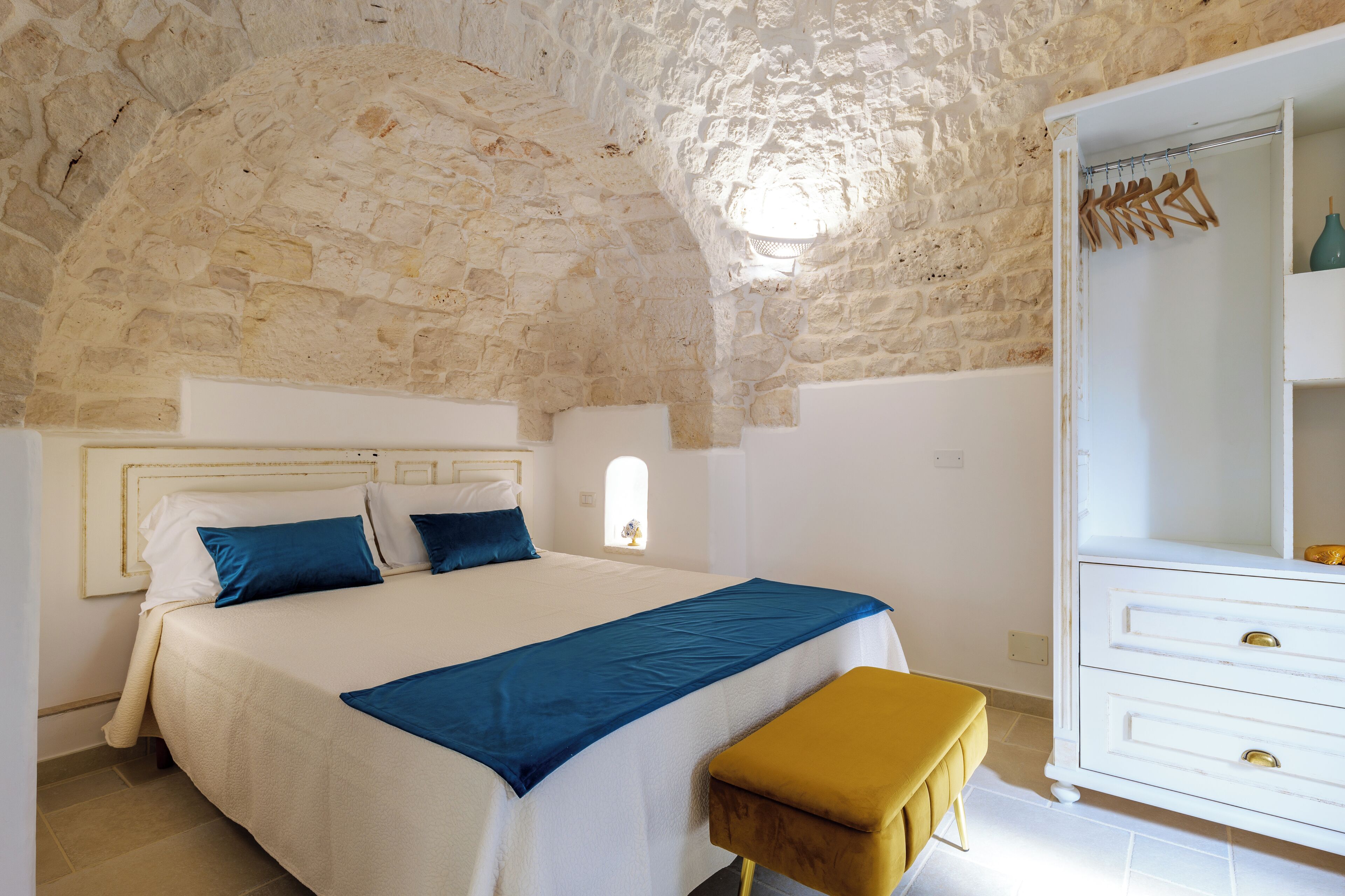 A bedroom with a stone ceiling, a bed with blue and white bedding, a wooden bench, and a white dresser with gold handles.