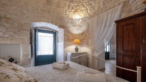 Quarto com parede de pedra, cama com roupa de cama floral, mesa de cabeceira com abajur e guarda-roupa de madeira.