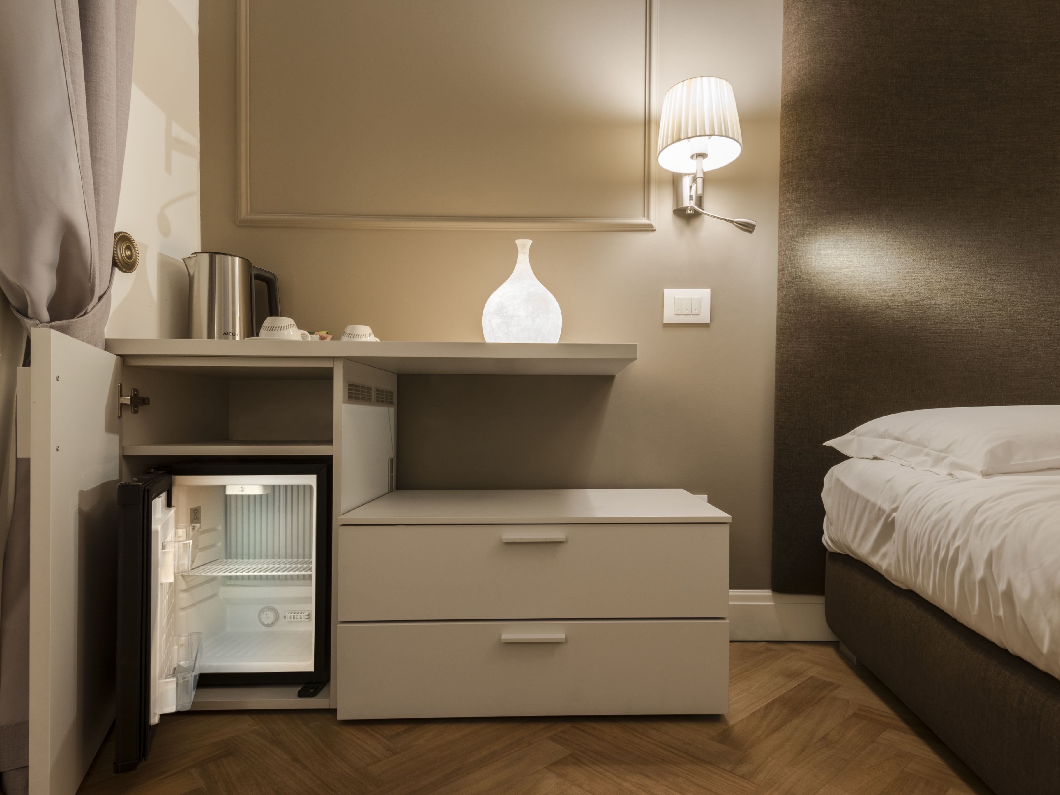 A compact hotel room with a bed, a bedside table with a lamp, a small refrigerator, and a built-in wardrobe.