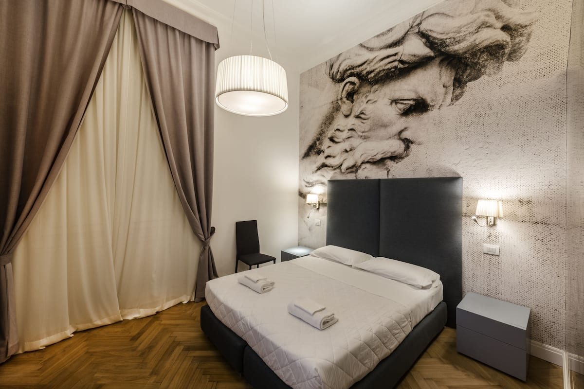 Una habitación de hotel moderna con una cama grande, cabecera, mesita de noche, una silla y una pared decorada con un cuadro abstracto de gran formato.