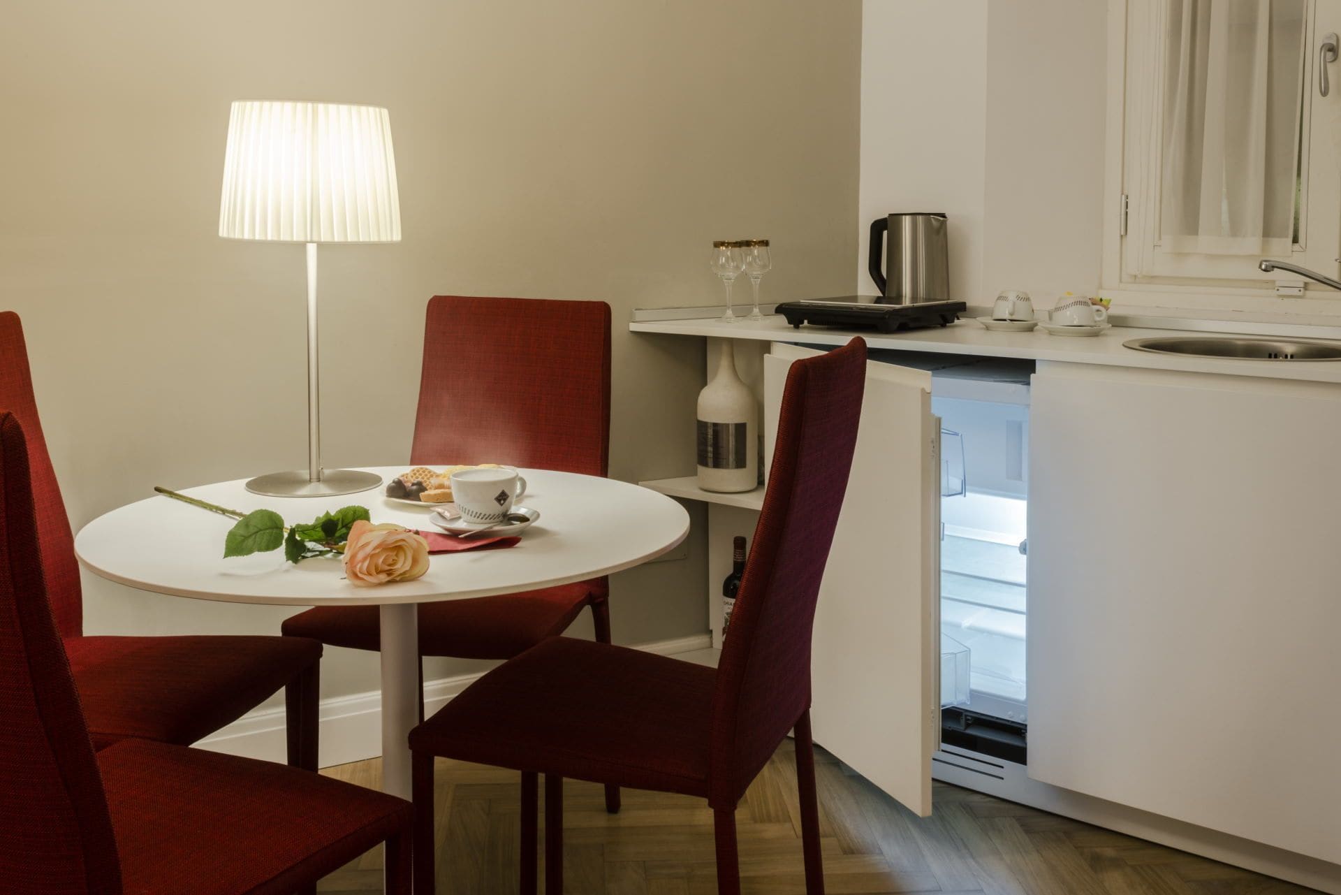 A compact kitchen with a dining area, including a table with a rose, a lamp, and a coffee cup.