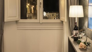 Una habitación de hotel con ventana que da a un paisaje urbano nocturno, una mesita de noche con una botella de vino, copas y una rosa.