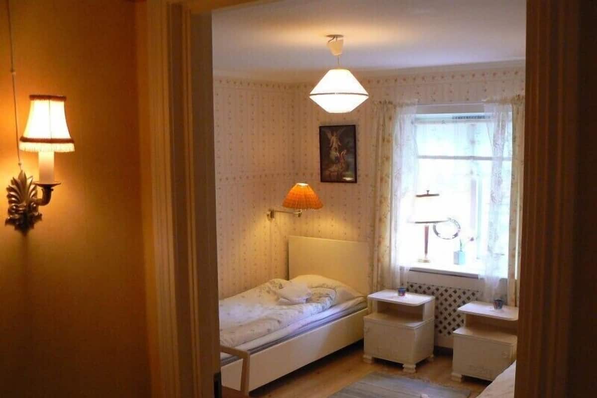 Une petite chambre à coucher accueillante, avec un lit simple, des tables de chevet, une commode, une fenêtre avec des rideaux et une lampe fixée au mur.
