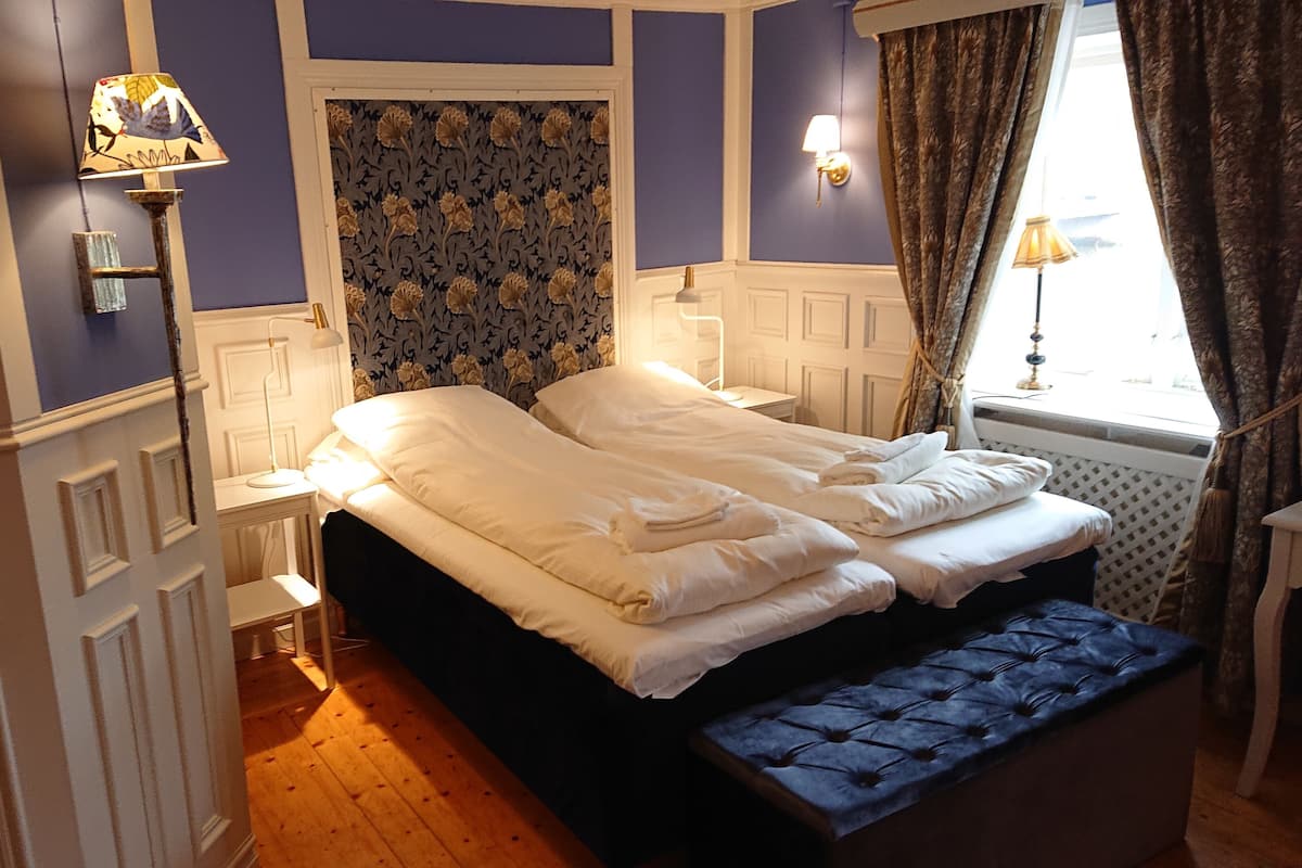 Une chambre à coucher avec un lit double, une banquette, une table de chevet, une lampe et une fenêtre avec des rideaux.