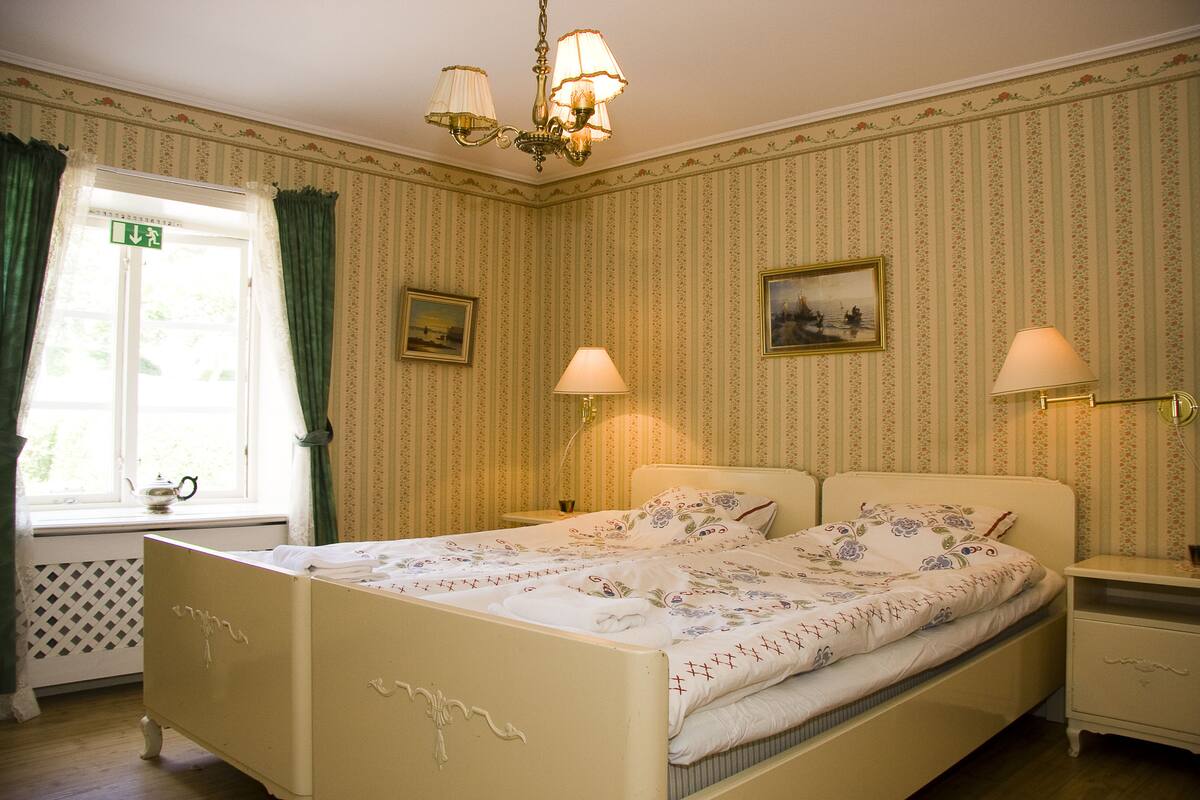 Une chambre à coucher avec un lit, deux tables de chevet, une fenêtre avec des rideaux et un lustre.