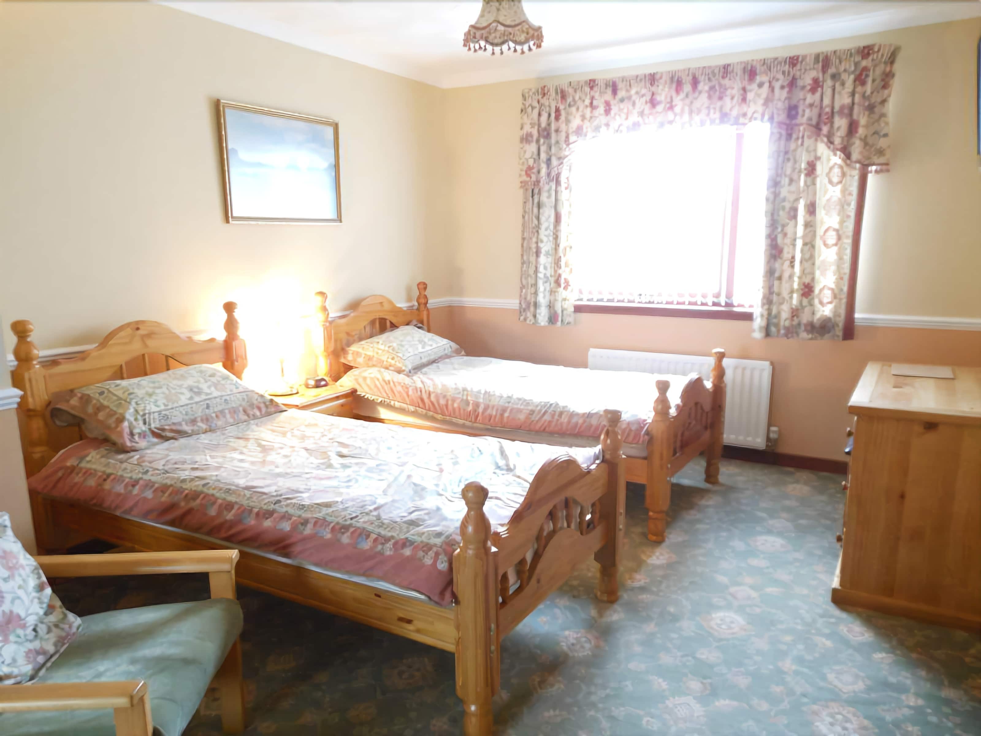 A bedroom with two wooden beds, a nightstand, a chair, a wooden cabinet, a radiator, and a window with floral curtains.