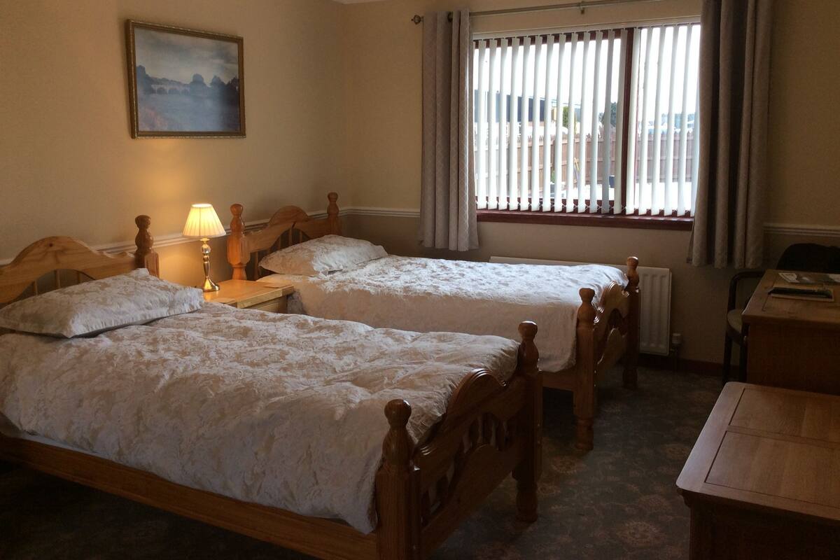 A room with two wooden beds, a nightstand with a lamp, a painting on the wall, and a window with blinds.