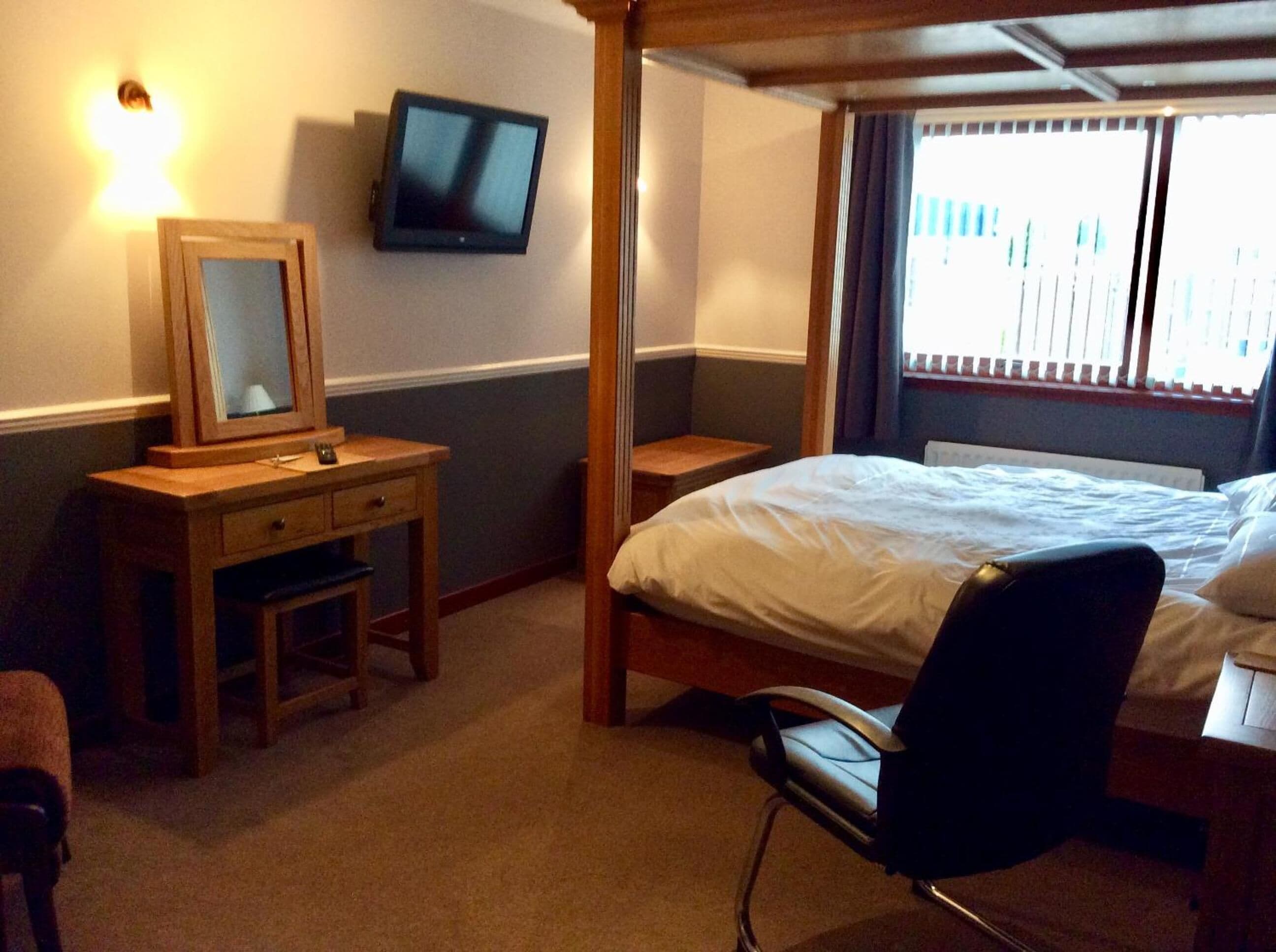 A bedroom with a bed, a desk with a mirror, a television on the wall, and a window with blinds.