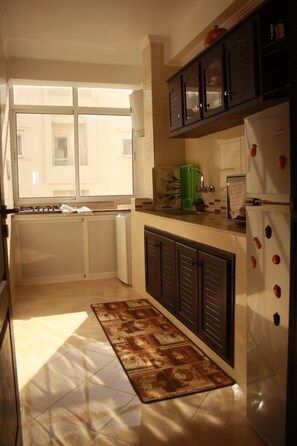 A kitchen with a window, cabinets, a refrigerator, and a patterned rug. - Coquets Appartements (Tangier)