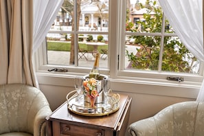 A room with a view of a garden through a window, a wooden side table with a tray holding a bottle and glasses, and two upholstered chairs.