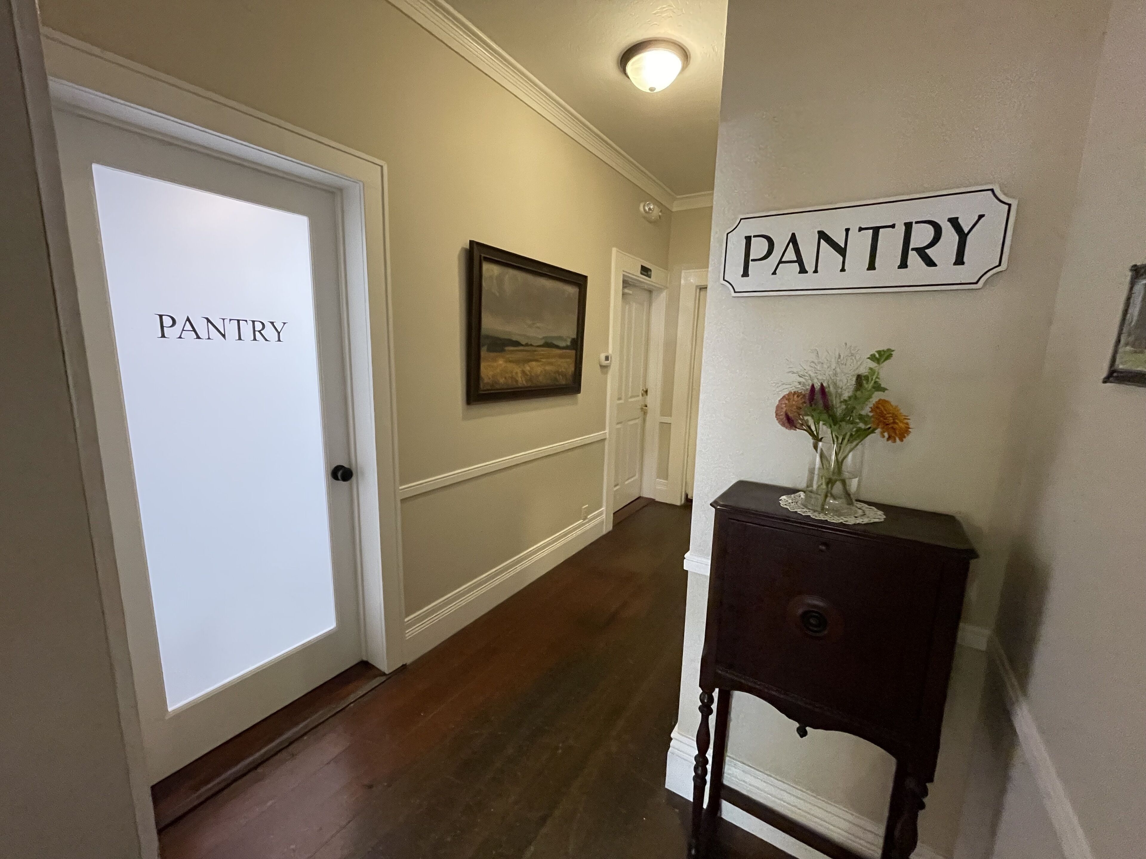 A hallway with a door labeled "PANTRY," a framed landscape painting, and a small table with a vase of flowers.