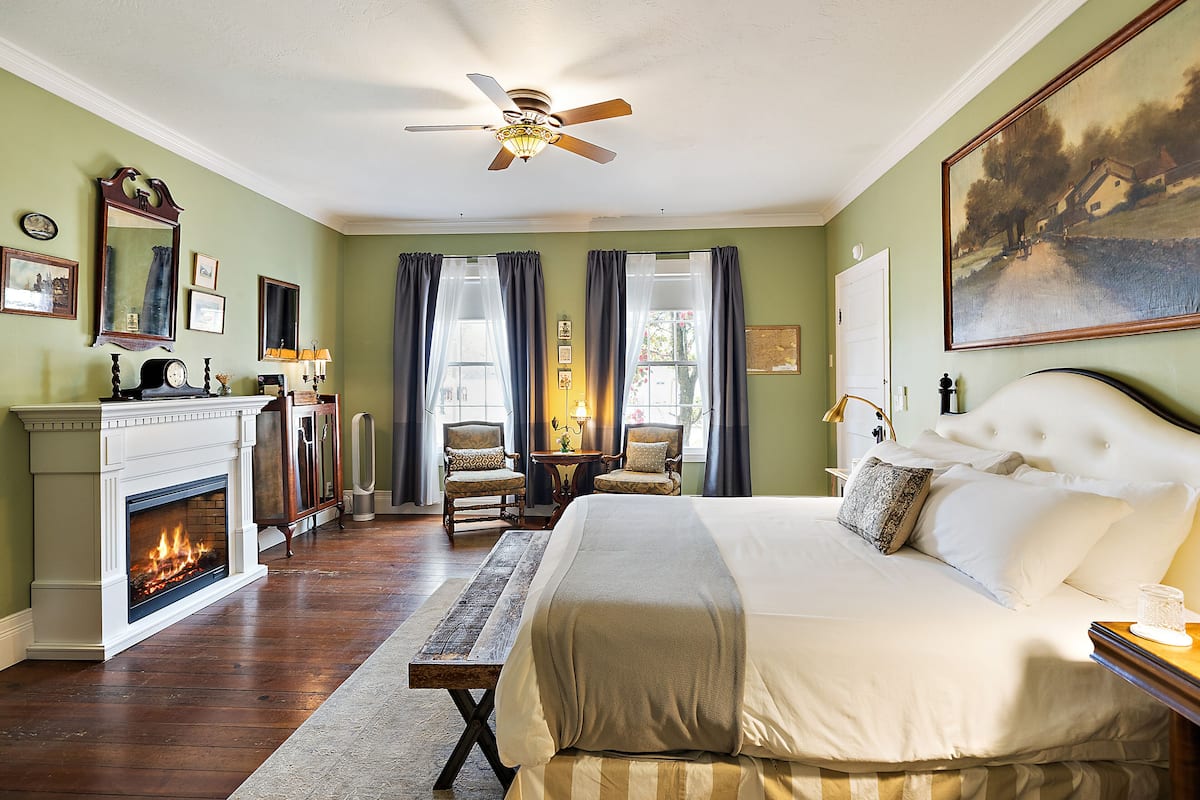 A bedroom with a fireplace, a large bed, a ceiling fan, and a painting on the wall.