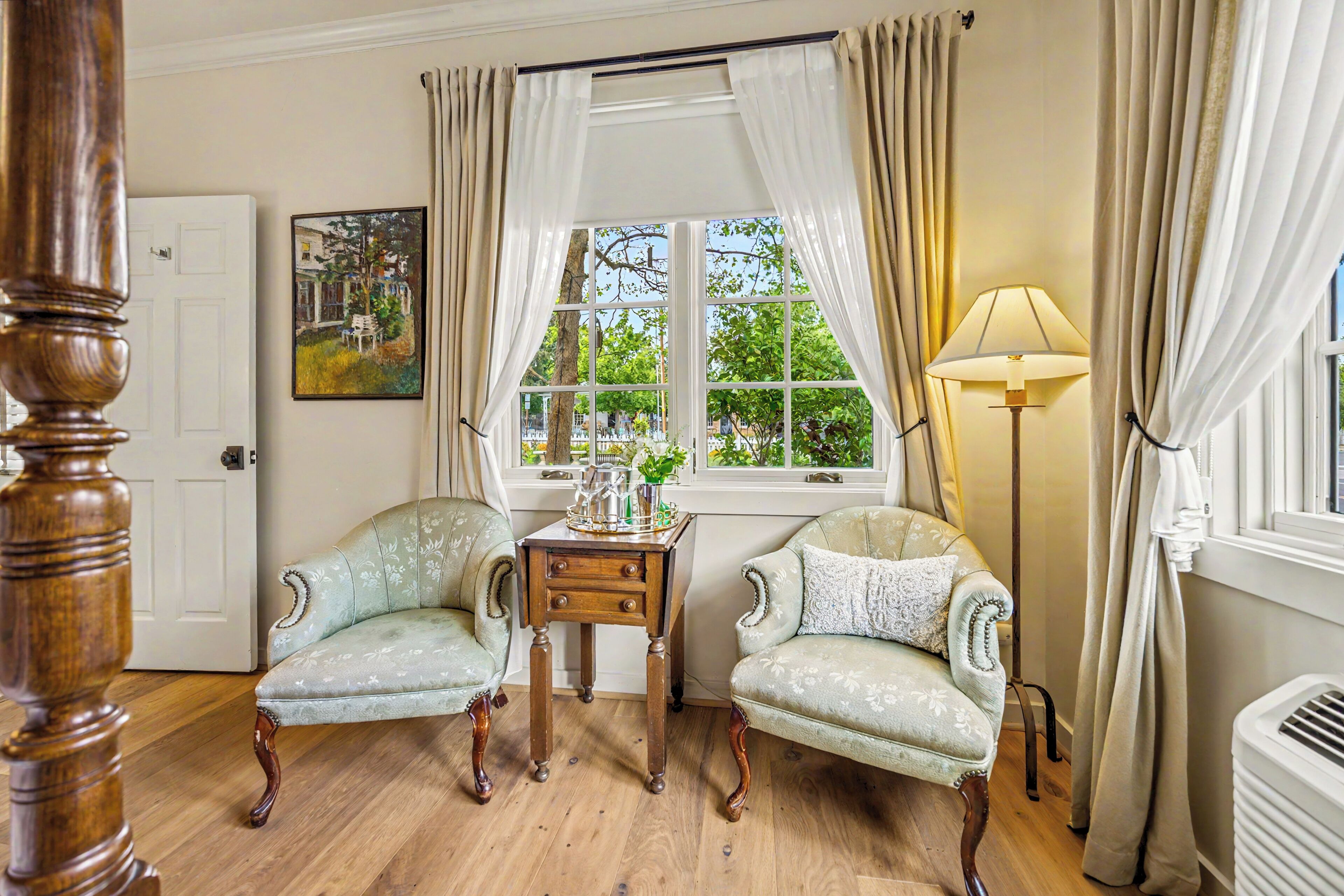 A room with two upholstered chairs, a small wooden table, a floor lamp, a window with curtains, and a painting on the wall.