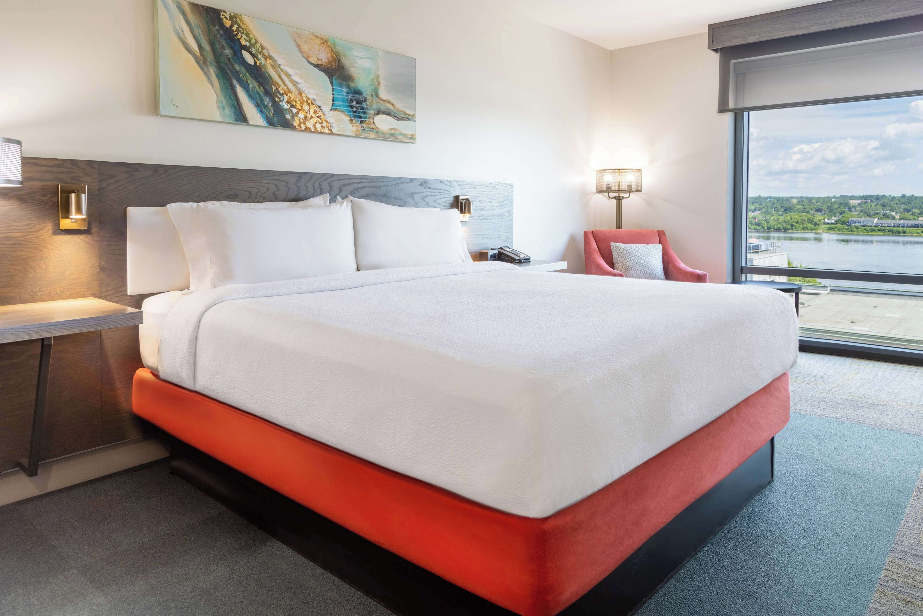 Habitación de hotel con una cama grande, una mesita de noche, una silla, un cuadro y vistas a una masa de agua.