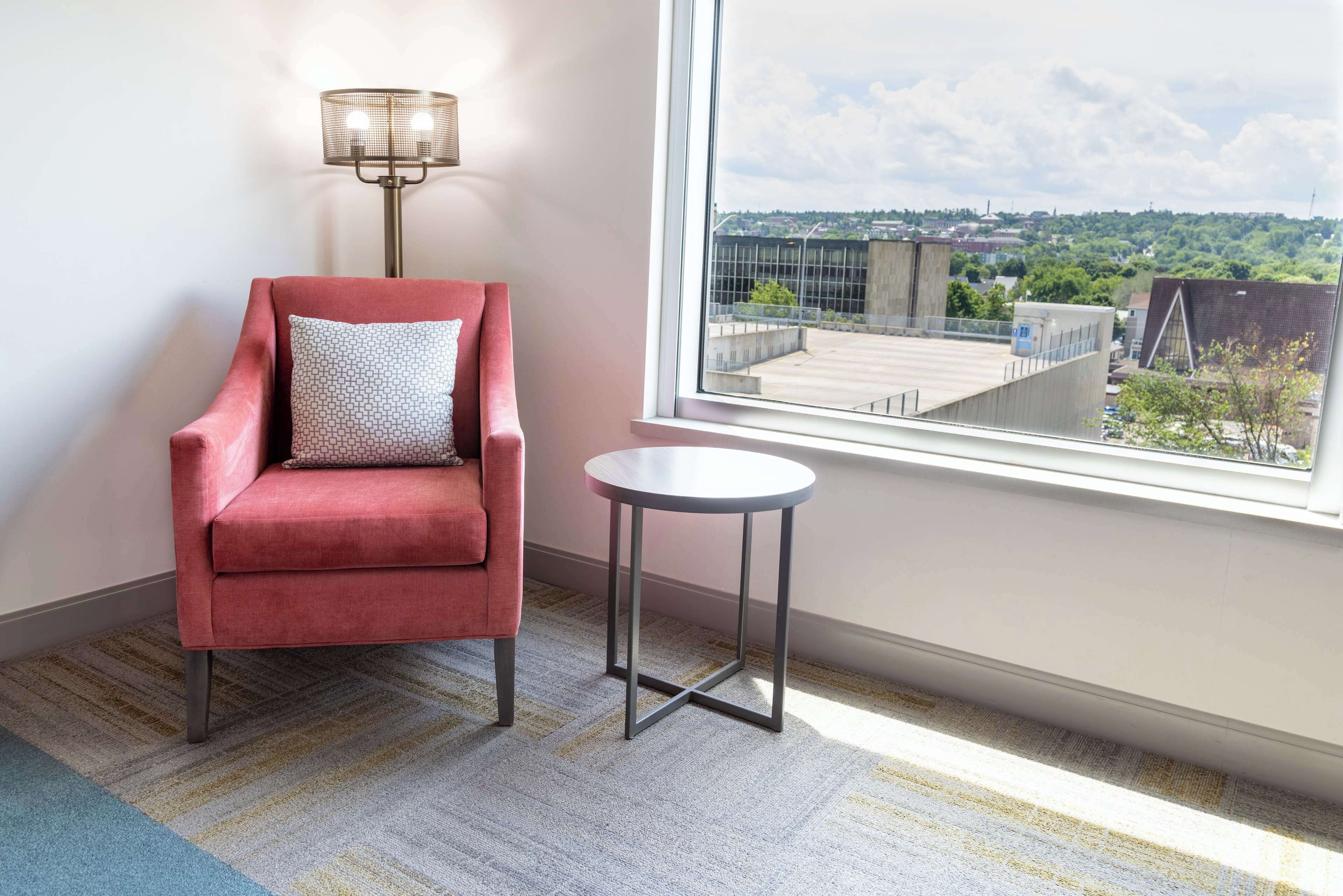 A red armchair with a patterned pillow, a small round table, and a view of a cityscape through a window.