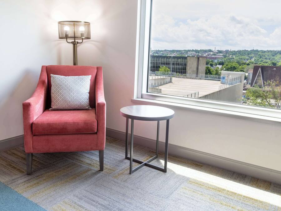 A red armchair with a patterned pillow, a small round table, and a view of a cityscape through a window.