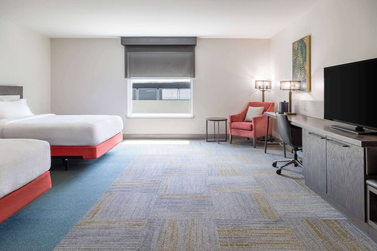 Une chambre d’hôtel avec deux lits, un bureau, une télévision et une fenêtre avec des stores.