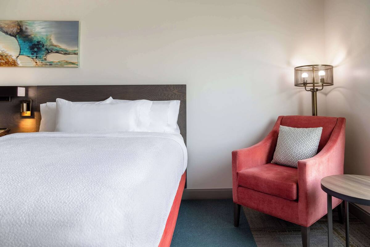 Une chambre d’hôtel avec un grand lit, un fauteuil rouge, une petite table et une lampe sur pied.