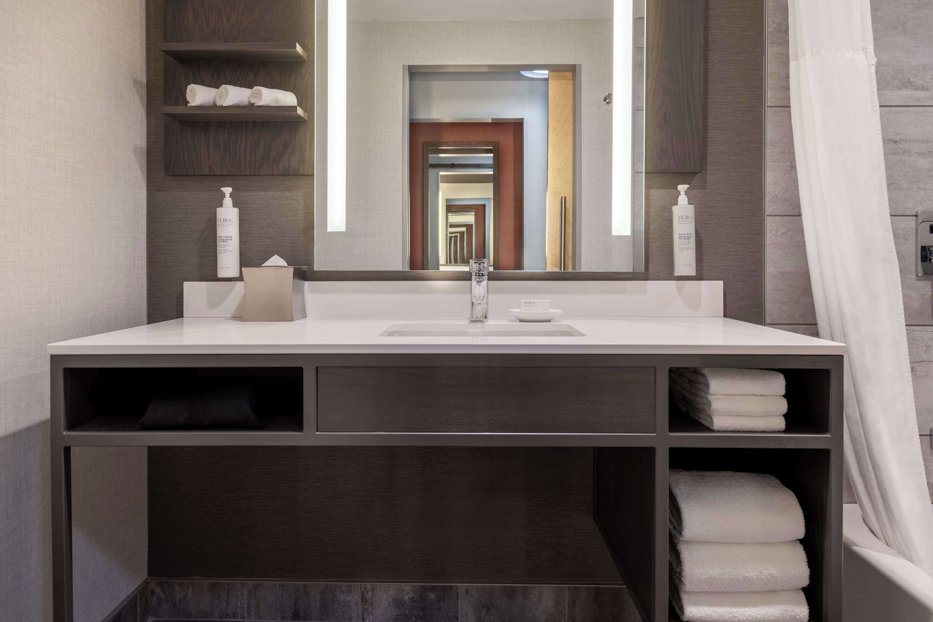 Un bagno moderno con uno specchio grande, lavandino bianco e scaffali a muro contenenti asciugamani e articoli da toilette.