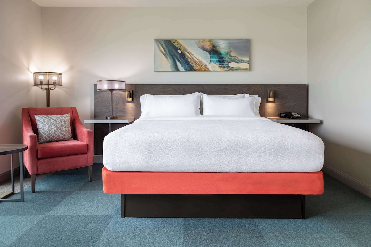 Une chambre d’hôtel avec un grand lit, un fauteuil rouge, une lampe sur pied et un tableau au mur.