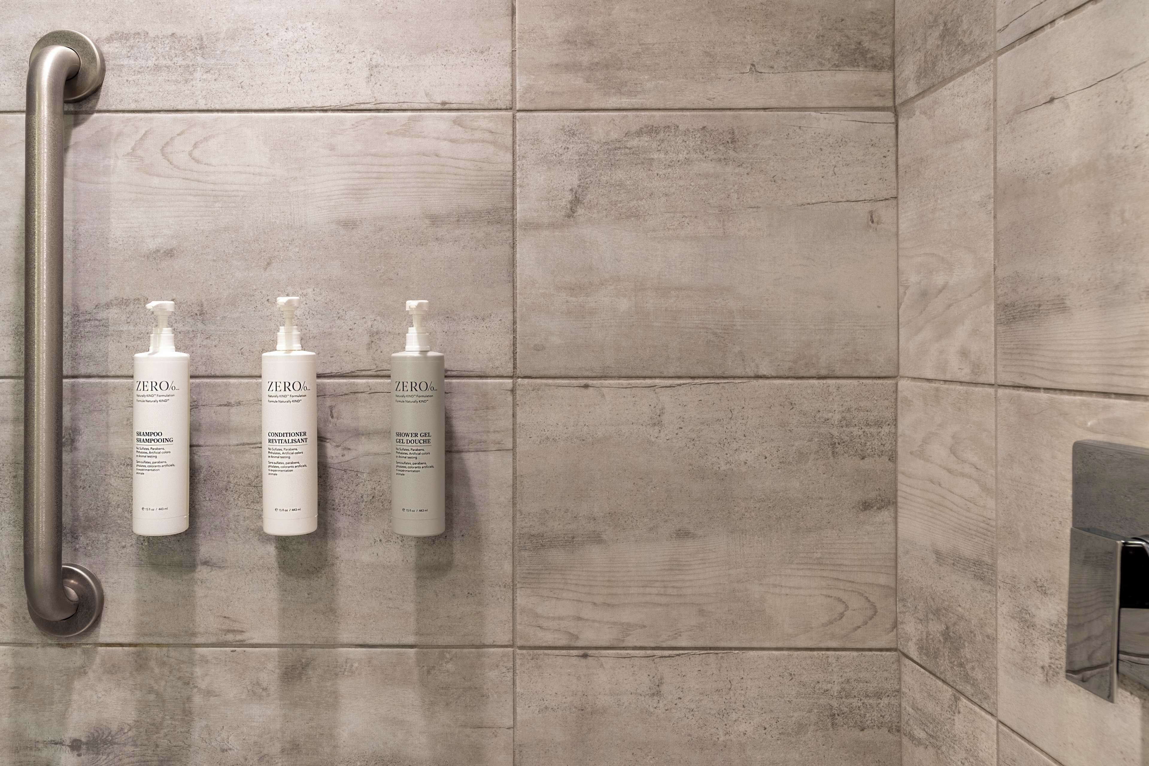 A bathroom with grey tiles, a handrail, and three bottles of 'ZERO%' shampoo, conditioner, and shower gel.