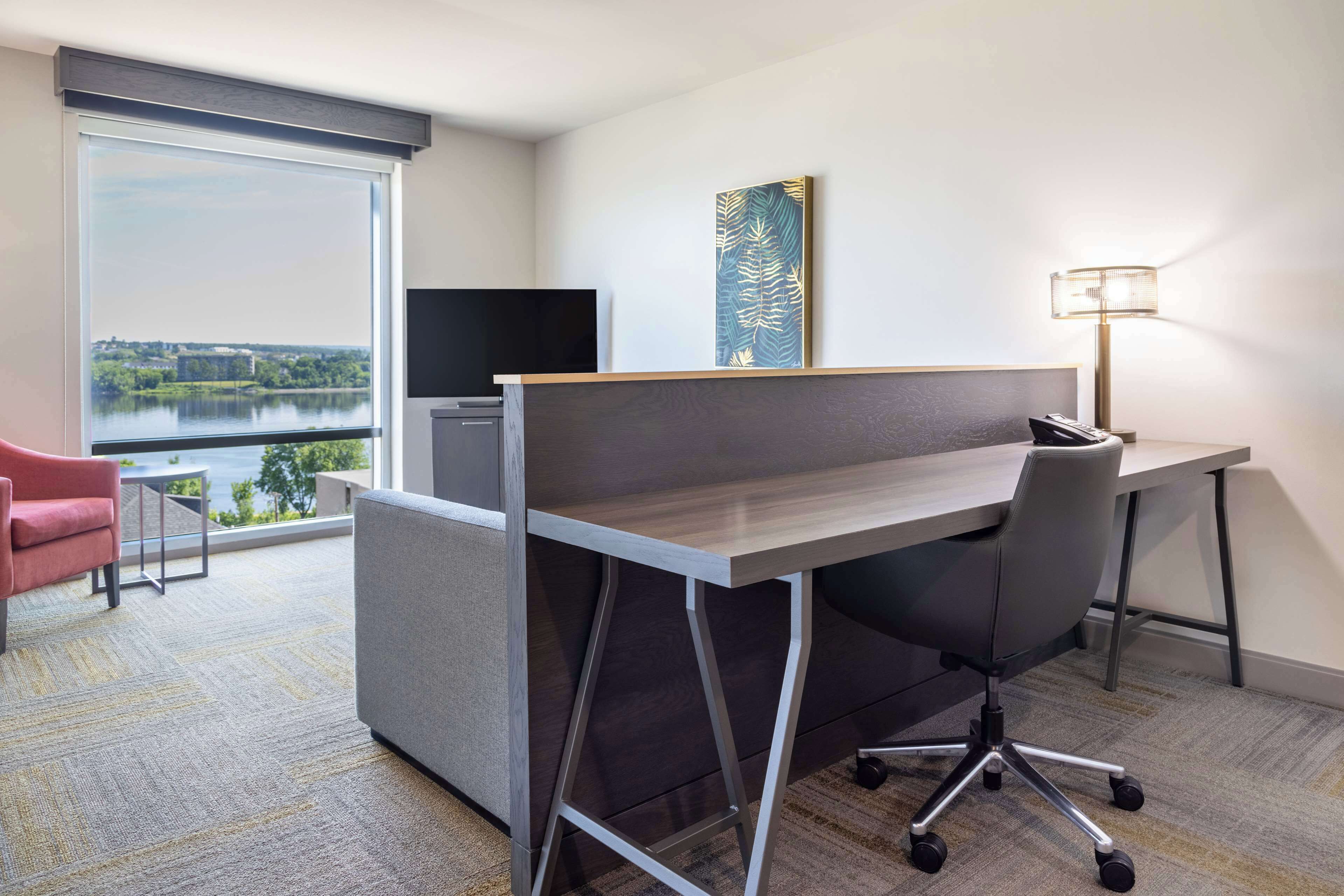 A modern hotel room with a large window offering a view of a lake and cityscape, a desk with a chair, a television, and a lamp.