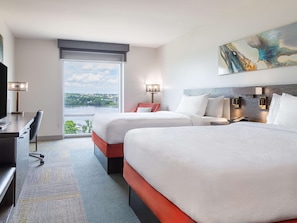 A hotel room with two beds, a desk, a chair, a TV, and a large window with a city view.