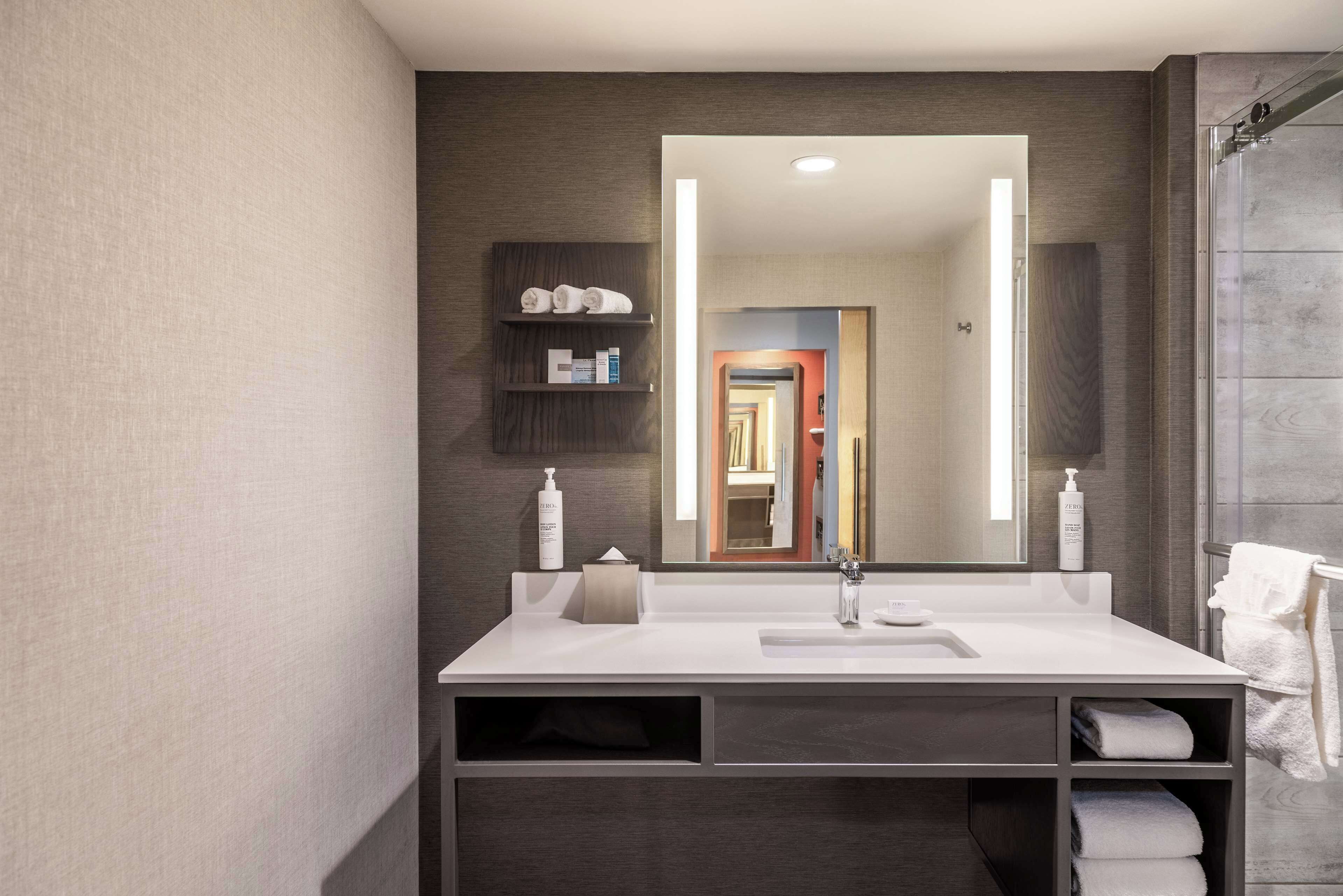 A modern bathroom with a large mirror, white sink, and dark wood shelving.