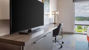 A hotel room with a large flat-screen TV, a desk with a chair, a lamp, and a view of a lake through a large window.