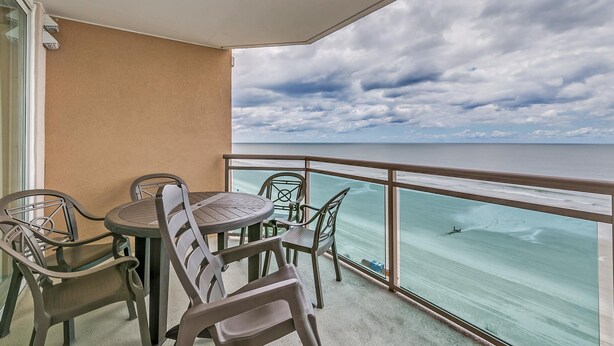 Un balcon avec une table et des chaises, donnant sur une plage à l’eau claire et sous un ciel nuageux.