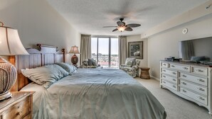 Un dormitorio con una cama grande, ventilador de techo, cómoda, televisor y vista a la ciudad desde la ventana.
