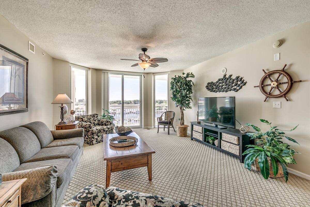 A spacious living room with a ceiling fan, large windows, a television, and a sail wheel decoration.