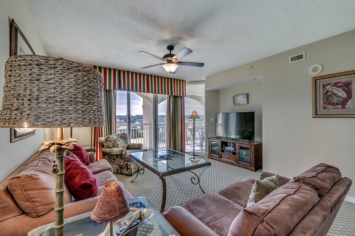 A living room with a brown leather sofa, a glass coffee table, a wicker lamp, a television, and a balcony with a view.