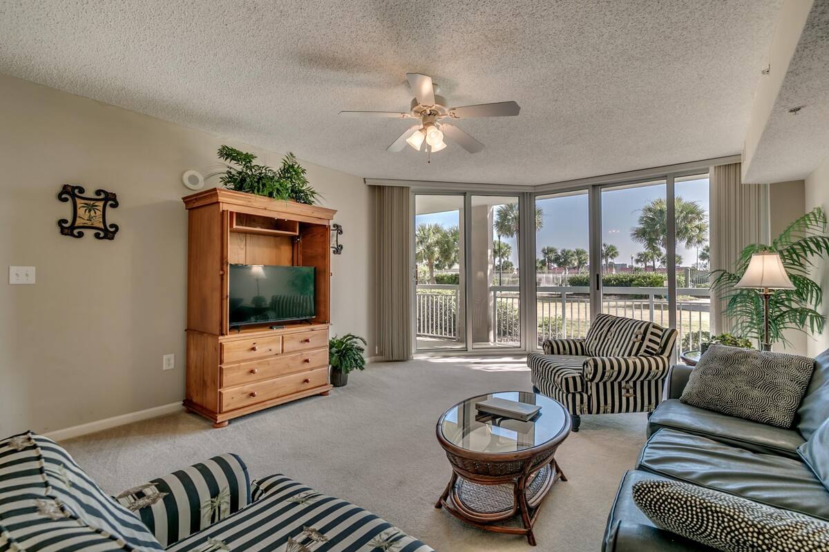 A spacious living room with a large flat-screen TV, a ceiling fan, and a view of palm trees through large sliding glass doors.