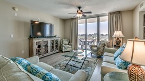 Una sala de estar moderna con una televisión de pantalla plana, asientos cómodos, una mesa de centro de vidrio y vistas de la ciudad a través de ventanas grandes.