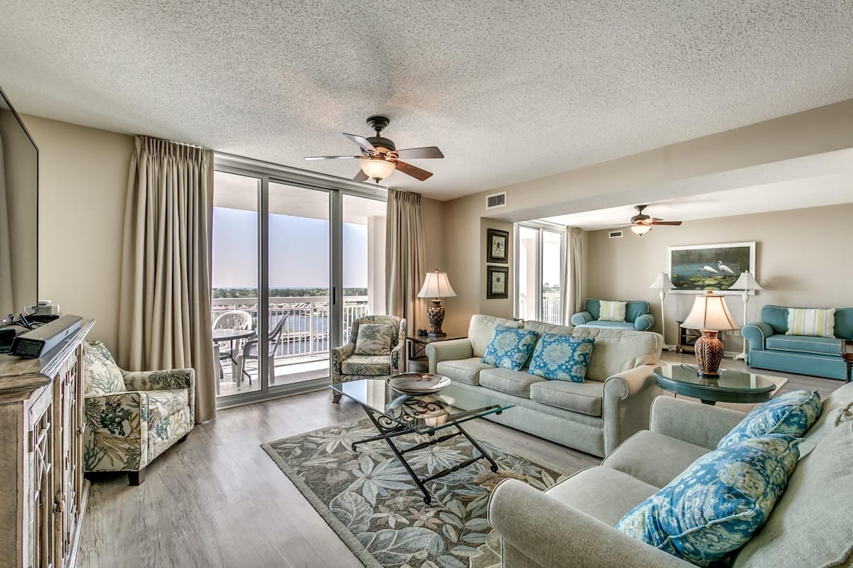A spacious living room with a large sliding glass door leading to a balcony, a ceiling fan, and a mix of seating options including a sofa and armchairs.