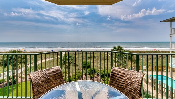 Ein Balkon mit Korbstühlen und einem Glastisch mit Blick auf einen Strand und den Ozean.
