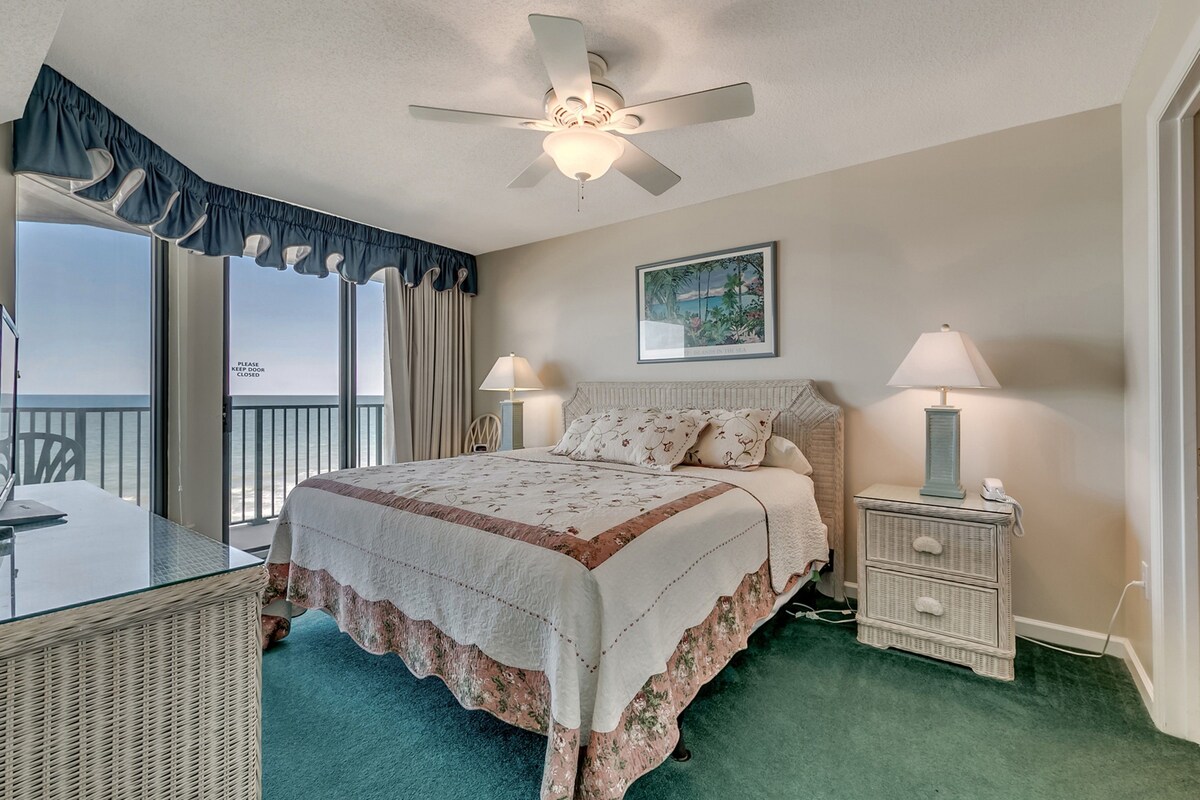A bedroom with a bed, two bedside tables with lamps, a ceiling fan, a window with curtains, and a view of the outdoors.