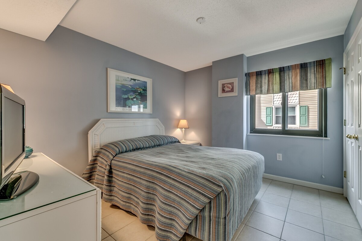 A bedroom with a bed, a bedside table, a lamp, a window with blinds, and a door.